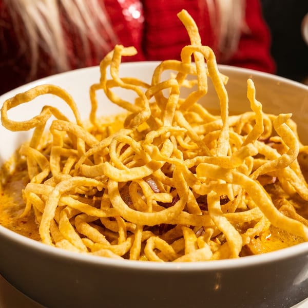 Close-up of a white bowl filled with golden crispy noodles and yellow curry, with a red sweater in the background.